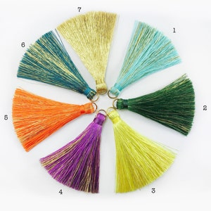 Peut inclure: Une collection de sept pompons colorés disposés en demi-cercle. Les pompons sont dans des tons de turquoise, vert, jaune, violet, orange et or. Chaque pompon est doté d'un anneau métallique doré. Les pompons sont faits d'une matière soyeuse.