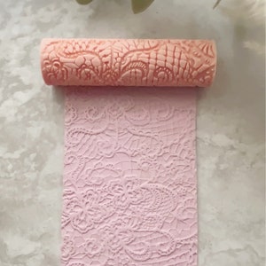 May include: A pink rolling pin with a lace pattern embossed on the surface. The rolling pin is laying on a piece of pink fondant with the same lace pattern embossed on it.