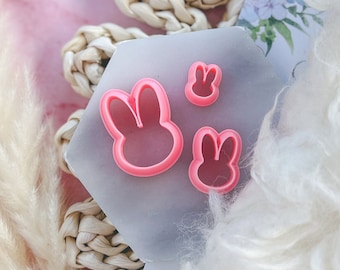May include: Three pink plastic bunny-shaped cookie cutters of different sizes on a gray surface.