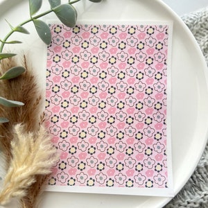 May include: A rectangular card with a pink floral pattern featuring light pink, yellow, gray, and black flowers. The card is displayed on a white plate, with greenery and pampas grass in the background.