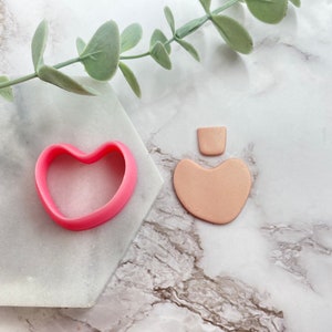 Op de afbeelding: Een roze hartvormige koekjesuitsteker en een lichtbruin hartvormig stuk fondant op een wit marmeren oppervlak.