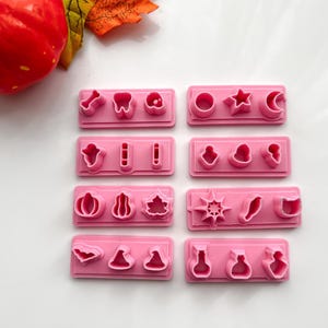 May include: Pink plastic cookie cutters in various shapes, including a tooth, star, and pumpkin. The cutters are arranged in rectangular sets. A red pumpkin and autumn leaves are in the background.