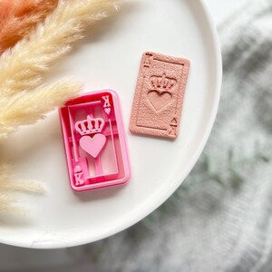 Puede incluir: Dos cortadores de galletas de plástico rosa con forma de cartas de juego. Los cortadores están en un plato blanco. La carta de la izquierda tiene un corazón y una corona. La carta de la derecha tiene un corazón y una pica.