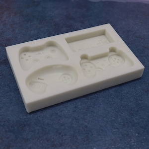 May include: White silicone mold with four different shapes of video game controllers. The mold is designed to create edible replicas of video game controllers.