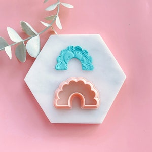 Puede incluir: Dos cortadores de galletas con forma de arcoíris, uno azul y otro rosa, sobre una superficie hexagonal blanca.