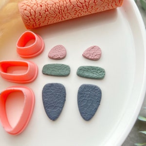 Op de afbeelding: Een set polymer clay oorbellen in verschillende kleuren en patronen. De oorbellen zijn in de vorm van druppels en rechthoeken. Er zijn ook drie roze plastic mallen voor het maken van oorbellen.