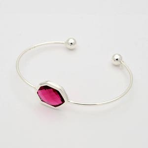 May include: A silver-toned cuff bracelet with a faceted, magenta-colored gemstone at the center. The bracelet has a simple, open design with a small silver ball at each end.