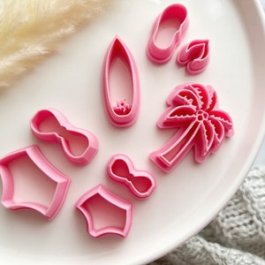 May include: A set of pink plastic cookie cutters shaped like a palm tree, flip flops, sunglasses, a surfboard, and a bow. The cutters are on a white plate.