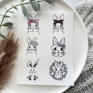 Pode incluir: Um papel branco com seis desenhos a traço preto de coelhos. Os coelhos são retratados com vários acessórios, incluindo bandanas florais, coroas de flores e gravatas borboleta. Os rostos dos coelhos são detalhados com bigodes e expressões.