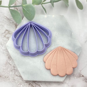 Puede incluir: Dos cortadores de galletas de concha de mar de plástico morado, uno es un contorno detallado de una concha de mar y el otro es una forma de concha de mar sólida.