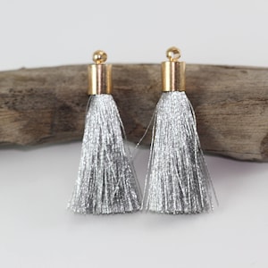 May include: A pair of silver tassel earrings. Each earring has a gold-coloured cap and a small gold bead. The tassels are made of shiny, metallic threads. The earrings are displayed on a white surface with driftwood in the background.