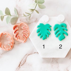 Puede incluir: Dos pares de pendientes de hoja de monstera verde en una pantalla hexagonal blanca con los números 1 y 2. Dos cortadores de galletas de hoja de monstera rosa están sobre una superficie de mármol blanco.