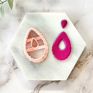 Puede incluir: Dos cortadores de galletas de plástico rosa, uno con forma de lágrima y el otro con forma de huevo con un corte en forma de lágrima. El cortador con forma de lágrima está relleno de fondant rosa.
