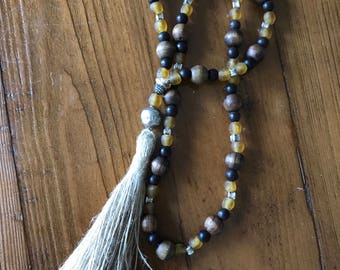 Op de afbeelding: Een kralenketting met een grote kwast. De ketting heeft houten kralen, zwarte kralen en gele kralen. De kwast is gemaakt van een lichtbruin materiaal. De ketting ligt op een houten oppervlak.