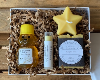 May include: A white gift box filled with brown shredded paper. The box contains a small bottle of honey, a lip balm, a bar of soap, and a yellow star-shaped candle. The honey bottle has a bear-shaped label with the text "B & G Honey Cato, NY 315-277-0538 Net Wt 1 oz". The lip balm label reads "B & G Honey Vanilla Bean Lip Balm Net Wt 1.5 oz". The soap label reads "B & G Honey Handmade Soap Pumpkin Spice Net Wt 3 oz".