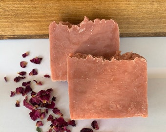 May include: Two bars of pink soap with a rough, textured edge are displayed. Scattered around the soap are dried red rose petals. The soap is a solid color, and the petals add a decorative element. The background is a light color with a wooden element.