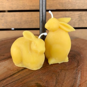 May include: Two yellow beeswax bunny candles on a wooden surface. The candles are shaped like bunnies and have white wicks.