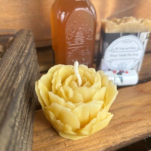 May include: A yellow beeswax candle shaped like a rose with a white wick. The candle is sitting on a wooden surface next to a glass jar of honey and a bar of soap.