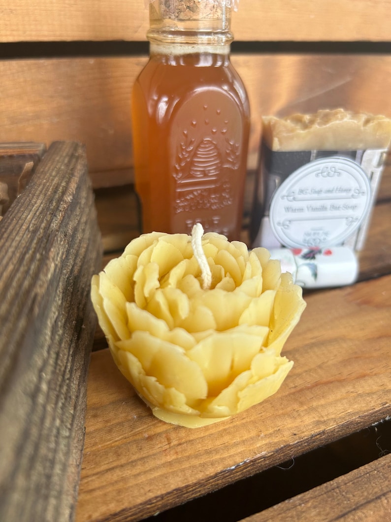 May include: A yellow beeswax candle shaped like a rose with a white wick. The candle is sitting on a wooden surface next to a glass jar of honey and a bar of soap.