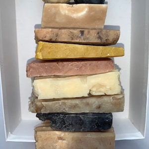 May include: A stack of handmade soap bars in a variety of colors and textures, including brown, beige, pink, yellow, and black. The soap bars are arranged in a white box.