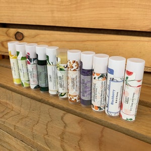 May include: A row of 12 lip balm tubes with various flavors, including Coconut, Vanilla Bean, Gingerbread, Lavender Lip Balm, Orange Creamsicle, Blackberry, and Cherry. Each tube has a white label with the flavor name and "B&G Soap and Honey" printed on it.