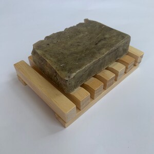 Puede incluir: Una barra de jab&oacute;n verde con motas marrones se encuentra sobre un jabonera de madera. La jabonera tiene un dise&ntilde;o de listones y est&aacute; hecha de madera clara.