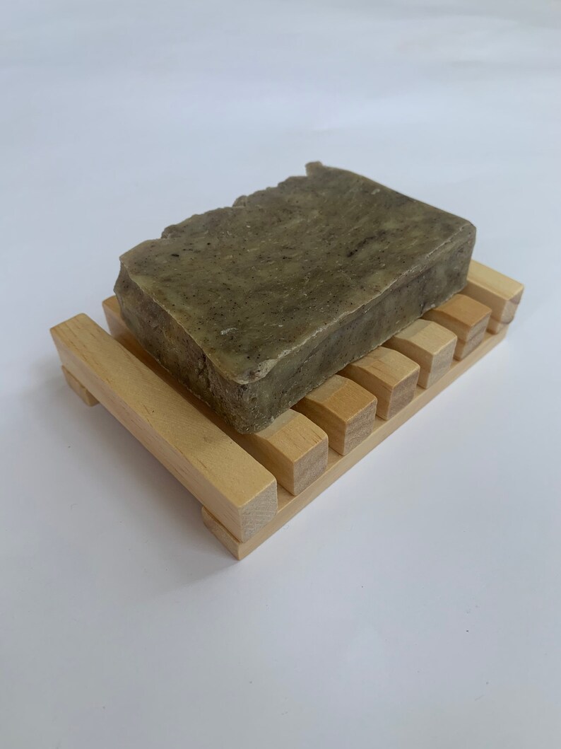 Puede incluir: Una barra de jab&oacute;n verde con motas marrones se encuentra sobre un jabonera de madera. La jabonera tiene un dise&ntilde;o de listones y est&aacute; hecha de madera clara.