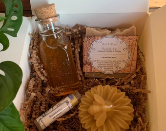 May include: A gift box filled with brown shredded paper, containing a small glass bottle of honey, a bar of oatmeal vanilla and honey soap, a small tube of blackberry lip balm, and a beeswax candle shaped like a bee.