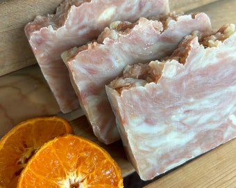 May include: Three bars of handmade soap with a swirl pattern in shades of brown and white. The soap is sitting on a wooden surface next to two orange slices.