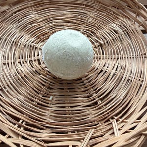 May include: A light green bath bomb sits in the center of a round, woven wicker basket. The bath bomb has a textured surface. The basket is made of thin, light brown woven strips.