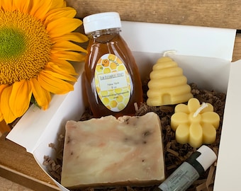 May include: A gift box containing a bottle of honey, a bar of soap, a lip balm, and two beeswax candles. The honey bottle is labeled "Raw Buckwheat Honey, New York." The lip balm is labeled "Peppermint, BG Soap and Honey." A large yellow sunflower is in the background.