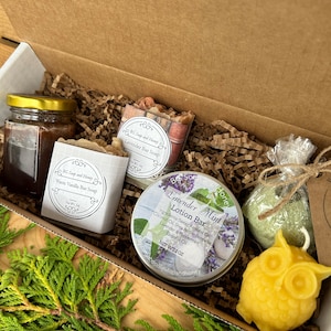 May include: A gift box filled with bath and body products. The box contains a jar of honey, a bar of warm vanilla soap, a bar of lavender soap, a tin of lavender mint lotion bar, a green bath bomb, and a yellow beeswax owl-shaped soap.
