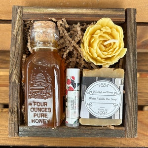 May include: A wooden gift box filled with a small bottle of honey, a lip balm, a bar of soap, and a yellow wax flower. The honey bottle is labeled "Four Ounces Pure Honey". The soap is labeled "BG Soap and Honey Warm Vanilla Bar Soap Net Wt. 2oz".