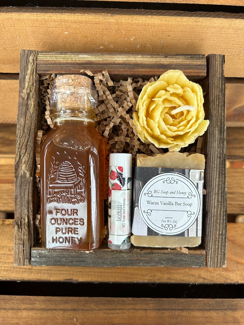 May include: A wooden gift box filled with a small bottle of honey, a lip balm, a bar of soap, and a yellow wax flower. The honey bottle is labeled "Four Ounces Pure Honey". The soap is labeled "BG Soap and Honey Warm Vanilla Bar Soap Net Wt. 2oz".