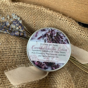 May include: A silver tin of Lavender Vanilla Lotion with a label that reads "B G Soap and Honey Lavender Vanilla Lotion" and lists the ingredients: Calendula Oil, Water Stearic Acid, Emulsifying Wax, Leucidal, Lavender and Vanilla Essential Oils. The label also states "NET WT 2 OZ" and "Etsy.com/shop/Bgsoapandhoney".