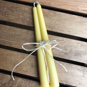 May include: Two yellow beeswax taper candles tied together with twine. The candles are on a wooden surface.