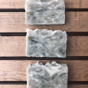 May include: Three rectangular bars of handmade soap with a marbled design. The soap features a blend of white and green hues, creating a natural, textured appearance. The bars are arranged on a wooden surface, showcasing their organic aesthetic.