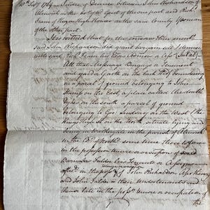 Puede incluir: Un documento antiguo de pergamino color hueso fechado el 10 de febrero de 1764, con texto manuscrito en tinta negra que detalla una escritura legal de traspaso entre John Richardson y Richard Fram, con pliegues horizontales visibles en el papel.