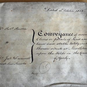 May include: A rectangular, aged parchment document dated 16th October 1812, featuring handwritten black ink text detailing a conveyance of land, barn, and stable in Middleton upon the Wolds, Yorkshire, between Mr. Robt. Raicton and Mr. Jwo. Dunwell.