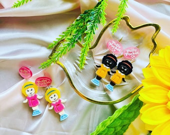 May include: Two pairs of earrings featuring Polly Pocket dolls. One pair features two black dolls wearing yellow dresses and the other pair features two blonde dolls wearing pink dresses. The earrings are on a white surface with green plants and a yellow flower.