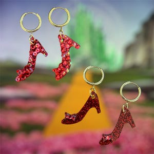 May include: Four pairs of red glitter high heel earrings with gold-colored hoops. The earrings are shaped like the iconic ruby slippers. The background features a yellow road and a green landscape.