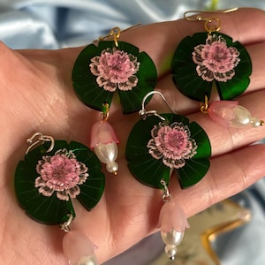 Peut inclure: Quatre paires de boucles d'oreilles avec un motif de nénuphar vert et une fleur rose au centre. Chaque boucle d'oreille a une fleur rose et une perle suspendues. Les boucles d'oreilles sont tenues dans une main.