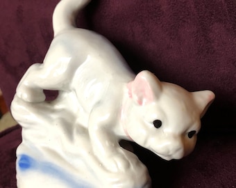 May include: White porcelain figurine of a cat with pink ears and black eyes, crouching on a blue and white base.