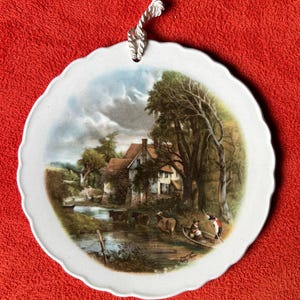 May include: A white ceramic plate with a scalloped edge, featuring a painted scene of a country cottage by a river with cows and people in a boat. The plate has a rope hanger.