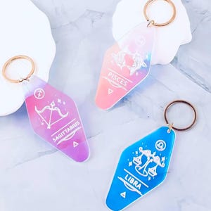 Może przedstawiać: Trzy iryzujące breloki do kluczy ze złotymi kółkami. Każdy brelok przedstawia inny znak zodiaku: Strzelca w kolorze fioletowym, Ryby w kolorze różowym i Wagę w kolorze niebieskim. Znaki zodiaku są wyświetlane z białą grafiką i tekstem.