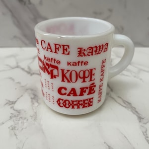 May include: White ceramic coffee mug with red text that reads "CAFE KAWA", "koffe", "KOFFE", "CAFE", and "COFFEE". The mug has a handle and is likely vintage.