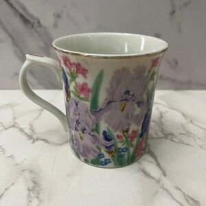 May include: White ceramic mug with a floral iris design in shades of purple, pink, and blue. The mug has a gold rim and handle. The flowers are set against a white background.