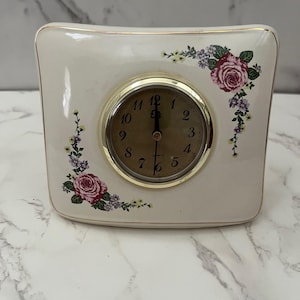 May include: Cream-colored ceramic quartz clock with a gold-tone bezel and a floral design. The clock face is tan with black numerals. Pink roses and purple flowers decorate the corners of the clock.