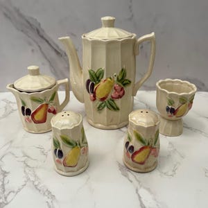 May include: A vintage cream-colored ceramic tea set featuring a teapot, sugar bowl, two salt shakers, and an egg cup. Each piece is decorated with a colorful fruit design including pears, plums, and cherries. The set has a ribbed design.