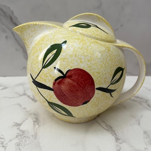May include: A vintage yellow ceramic pitcher with a speckled finish and a lid. The pitcher features a red apple design with green leaves and black stems. It has a curved handle and a spout for pouring.